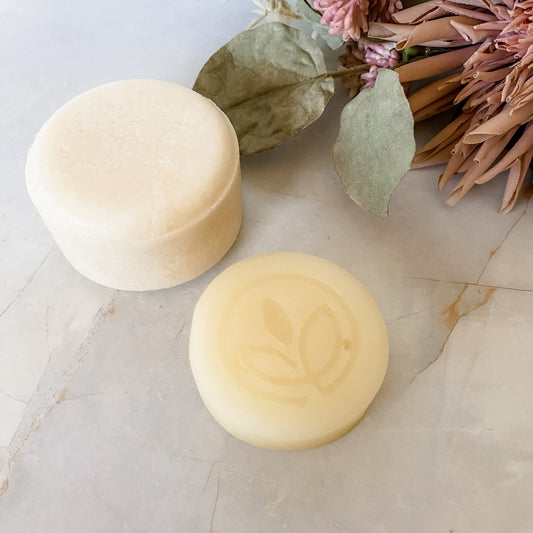 Plain cream shampoo and logo imprinted conditioner bars on a light marble surface with leaves & flowers in the background