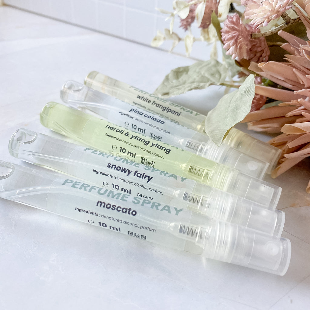 Five clear perfume spray vials with different colored liquids, labeled with scents including White Fragrance, Pina Colada, Neroli & Ylang Ylang, Snowy Fairy, and Moscato, arranged on a white surface with dried flowers in the background.