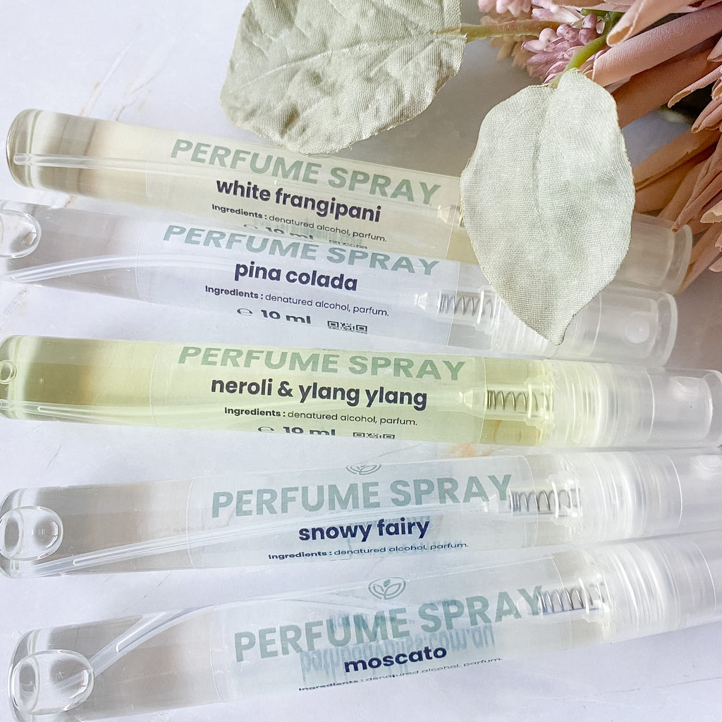 Five perfume spray tubes arranged diagonally on a white surface with decorative dried leaves and pink flowers. The tubes are clear and show pale yellow to off-white liquid inside, with printed labels for scents including white frangipani, pina colada, neroli & ylang ylang, snowy fairy, and moscato.