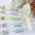 Five perfume spray tubes arranged diagonally on a white surface with decorative dried leaves and pink flowers. The tubes are clear and show pale yellow to off-white liquid inside, with printed labels for scents including white frangipani, pina colada, neroli & ylang ylang, snowy fairy, and moscato.