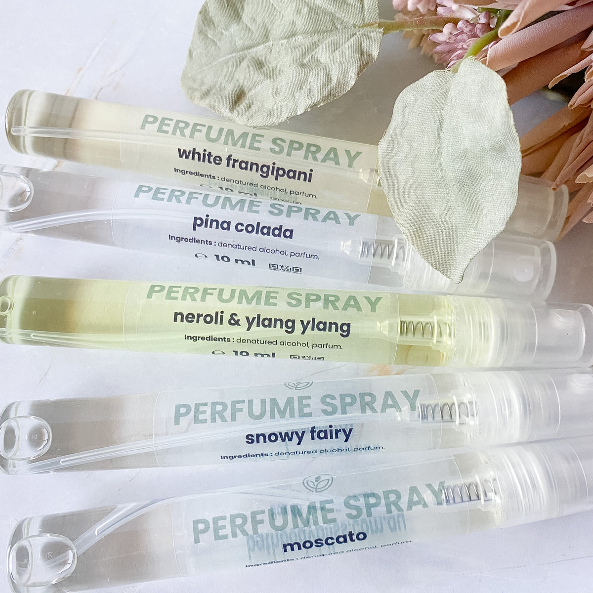 Five perfume spray tubes arranged diagonally on a white surface with decorative dried leaves and pink flowers. The tubes are clear and show pale yellow to off-white liquid inside, with printed labels for scents including white frangipani, pina colada, neroli & ylang ylang, snowy fairy, and moscato.