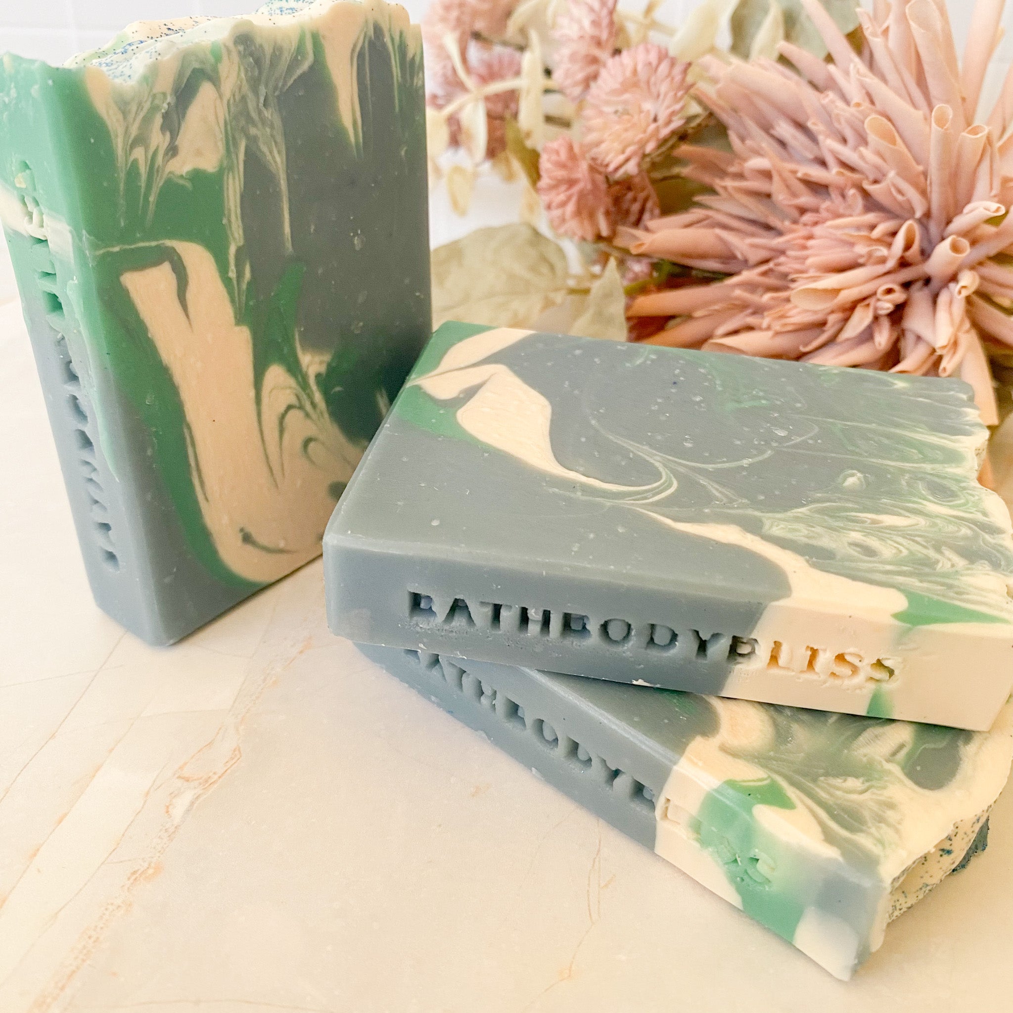 three bars of Bath Body Bliss soap with marbled design on a light surface with flowers in the background.