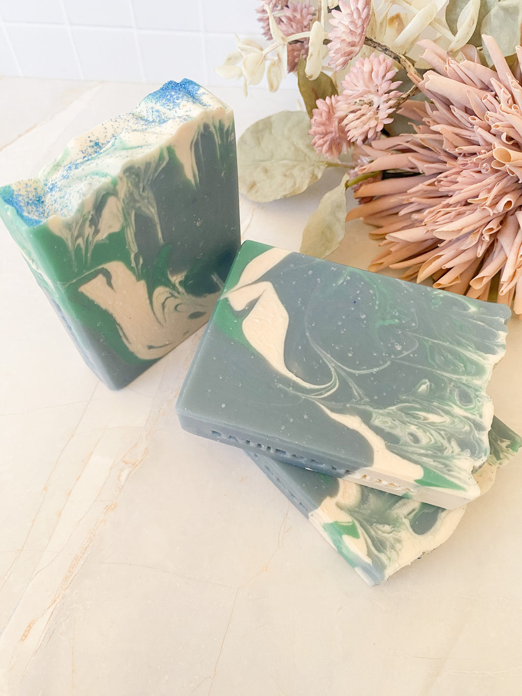 three bars of soap with green and white marbled patterns on a light surface with flowers in the background.