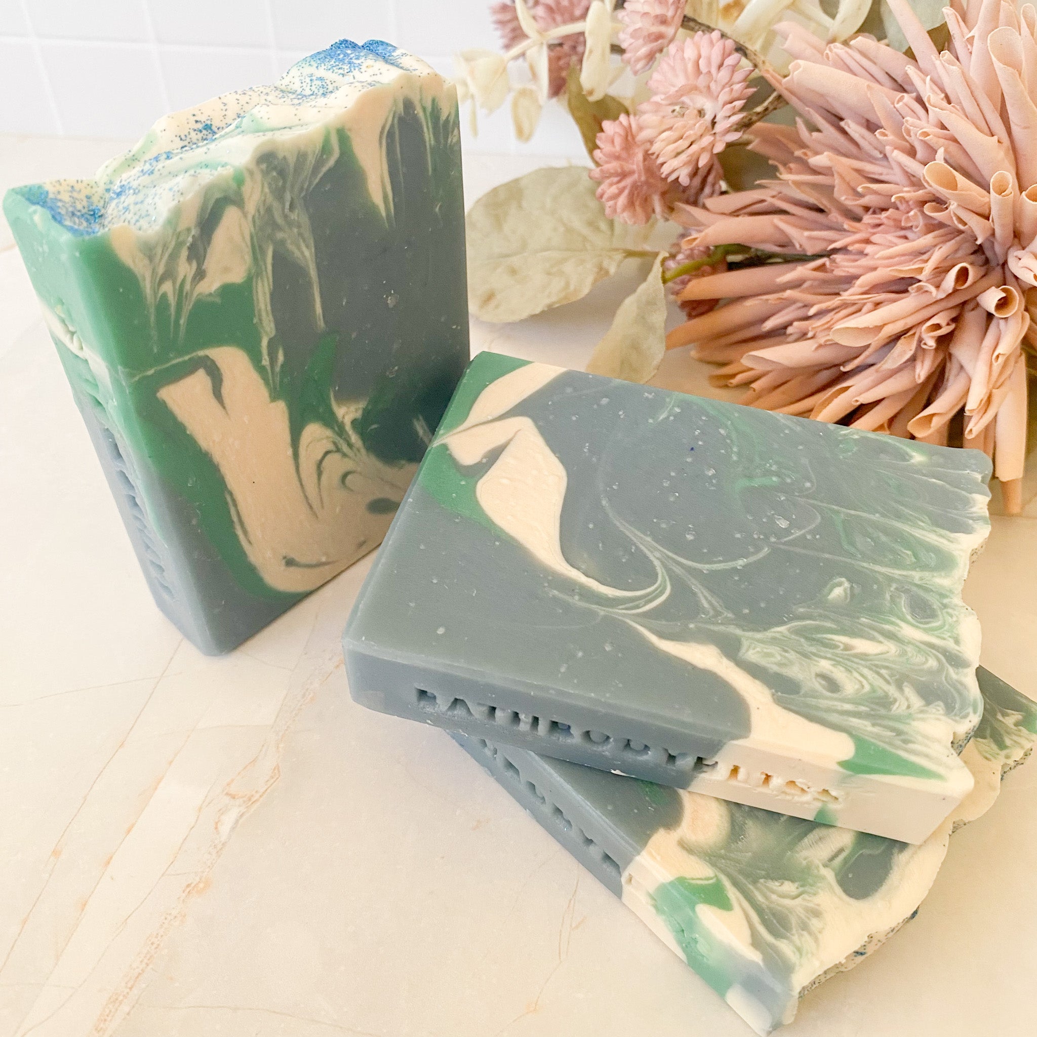 Marbled green and beige soap bars on a light surface with flowers in the background