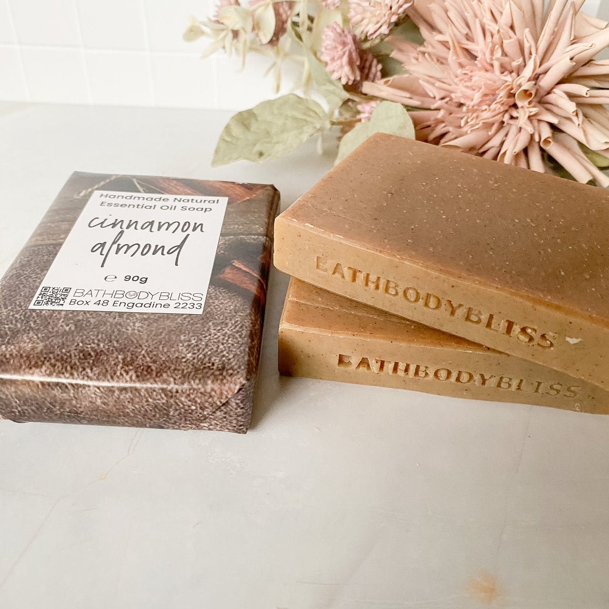 Three bars of Bathbody Bliss essential oil soap, two unwrapped showing cinnamon almond ingredients and one wrapped, displayed next to a matching wrapped bar on a light marble surface with dried flowers in the background.