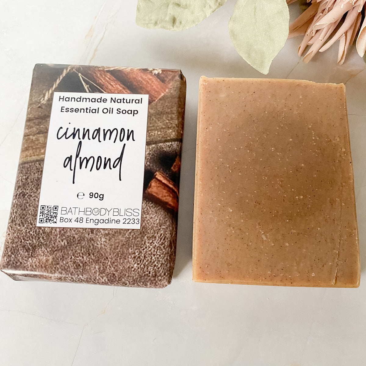 A bar of handmade essential oil soap next to an unwrapped bar. The soap is a light brownish-beige color with visible almond meal embedded in it, and the paper wrapped bar is labeled 'cinnamon almond' by Bath Body Bliss.