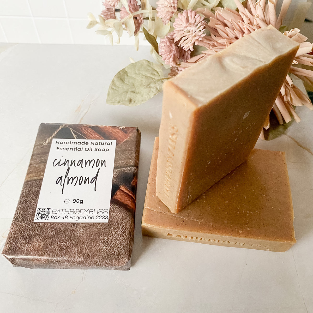three bars of handmade cinnamon almond soap, one wrapped in paper packaging, and two stacked beside it with floral decor in the background