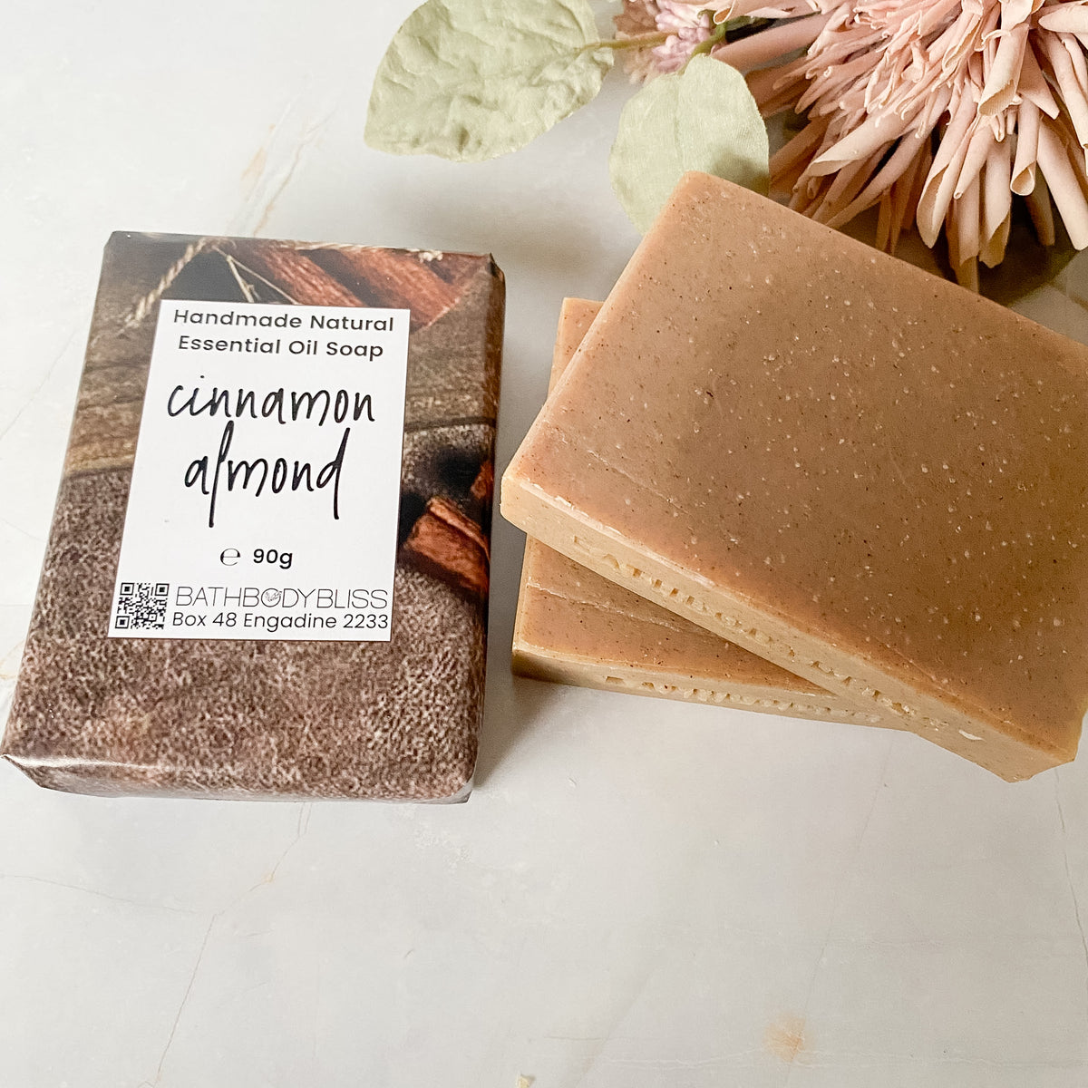 A 90g bar of Bath Body Bliss 'cinnamon almond' essential oil soap shown next to its two unwrapped bars on a light marble surface with dried flowers in the background.