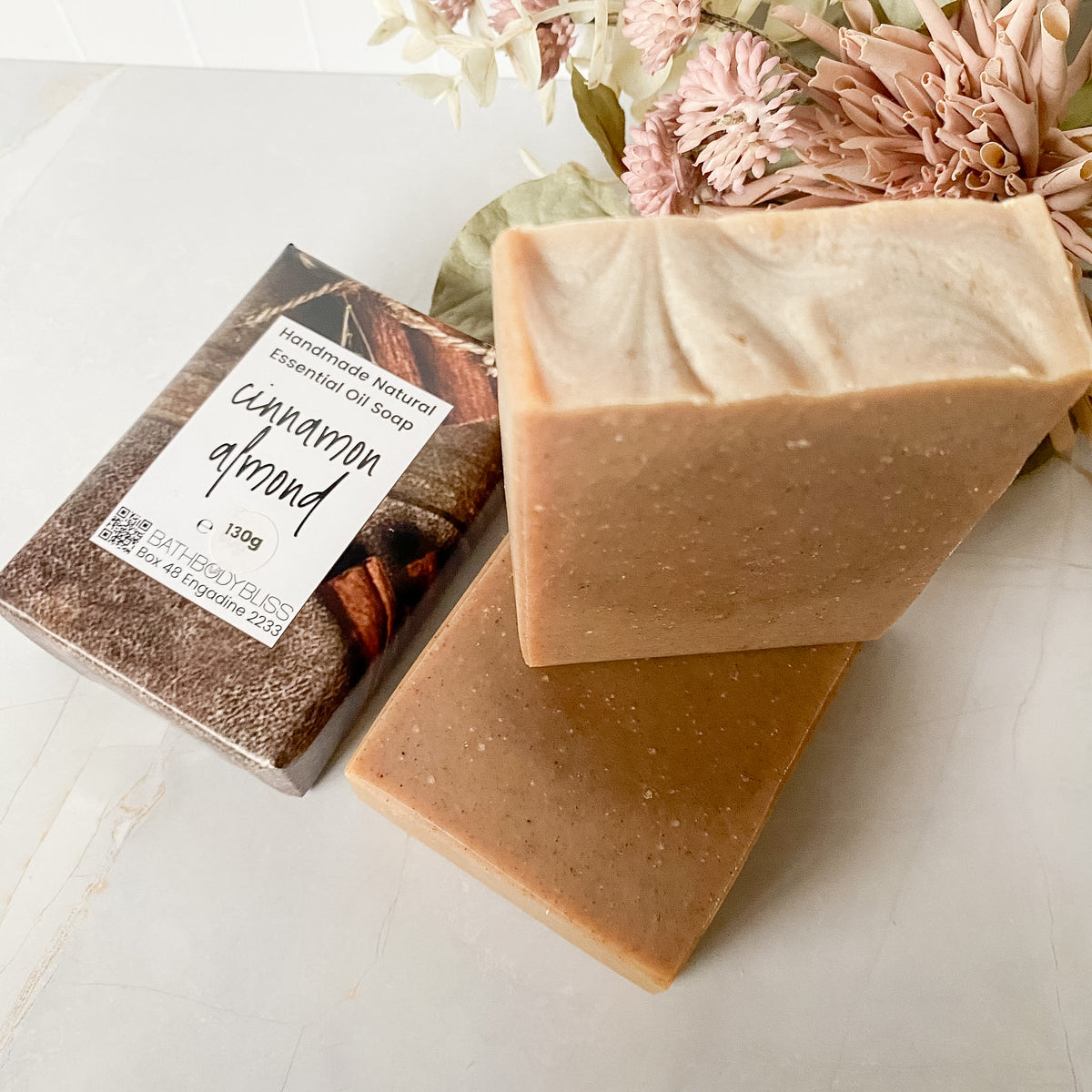 Two rectangular bars of handmade natural essential oil soap in warm beige and caramel-brown shades, with one bar stacked on top of the other. A wrapped bar labeled 'cinnamon almond' sits beside them on a light marble-like surface with dried pink flowers in the background.