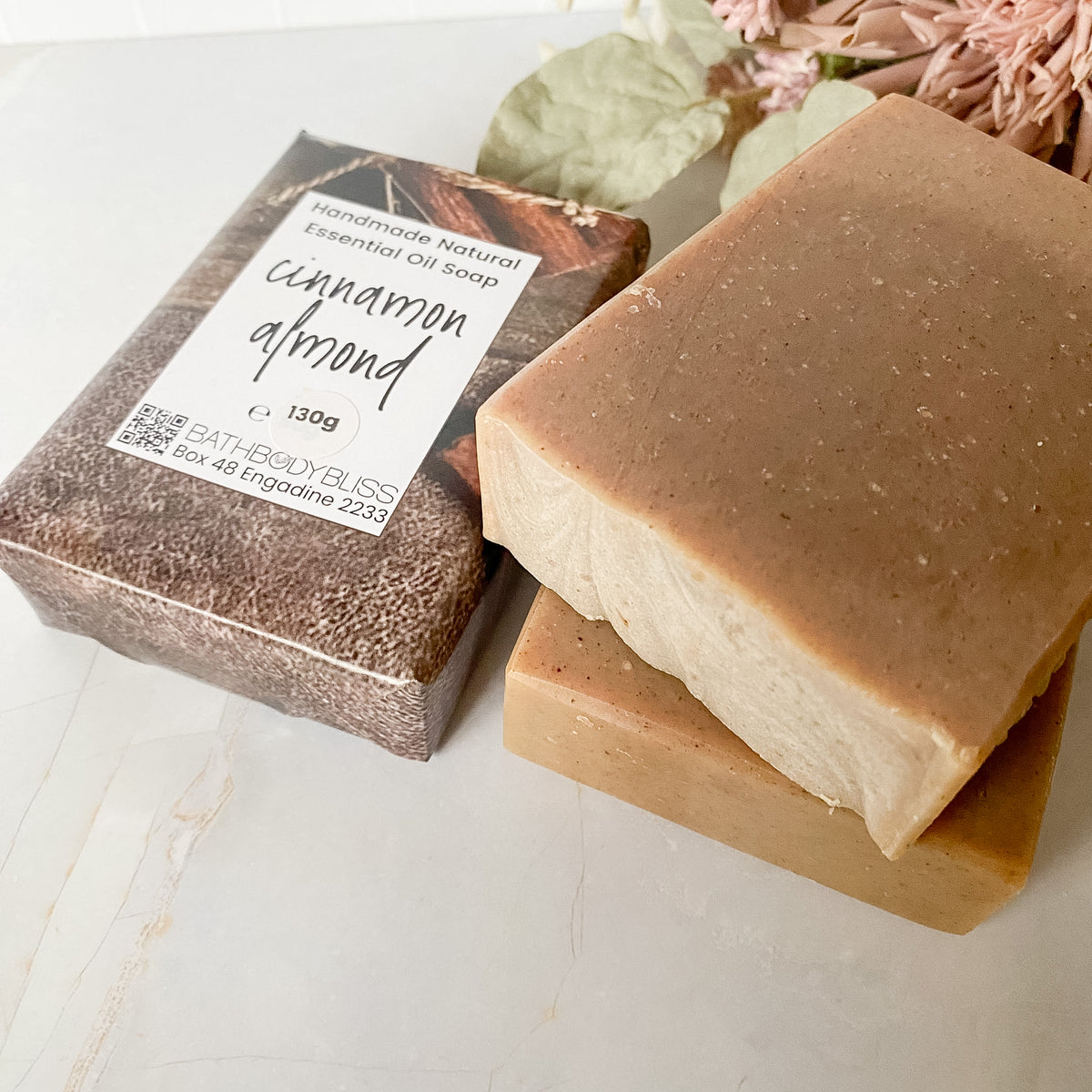 Three bars of Bath Body Bliss essential oil soap, one wrapped in brown paper with a white label reading 'cinnamon almond' and two unwrapped showing a warm tan surface, styled on a light marble surface with dried flowers in the background.