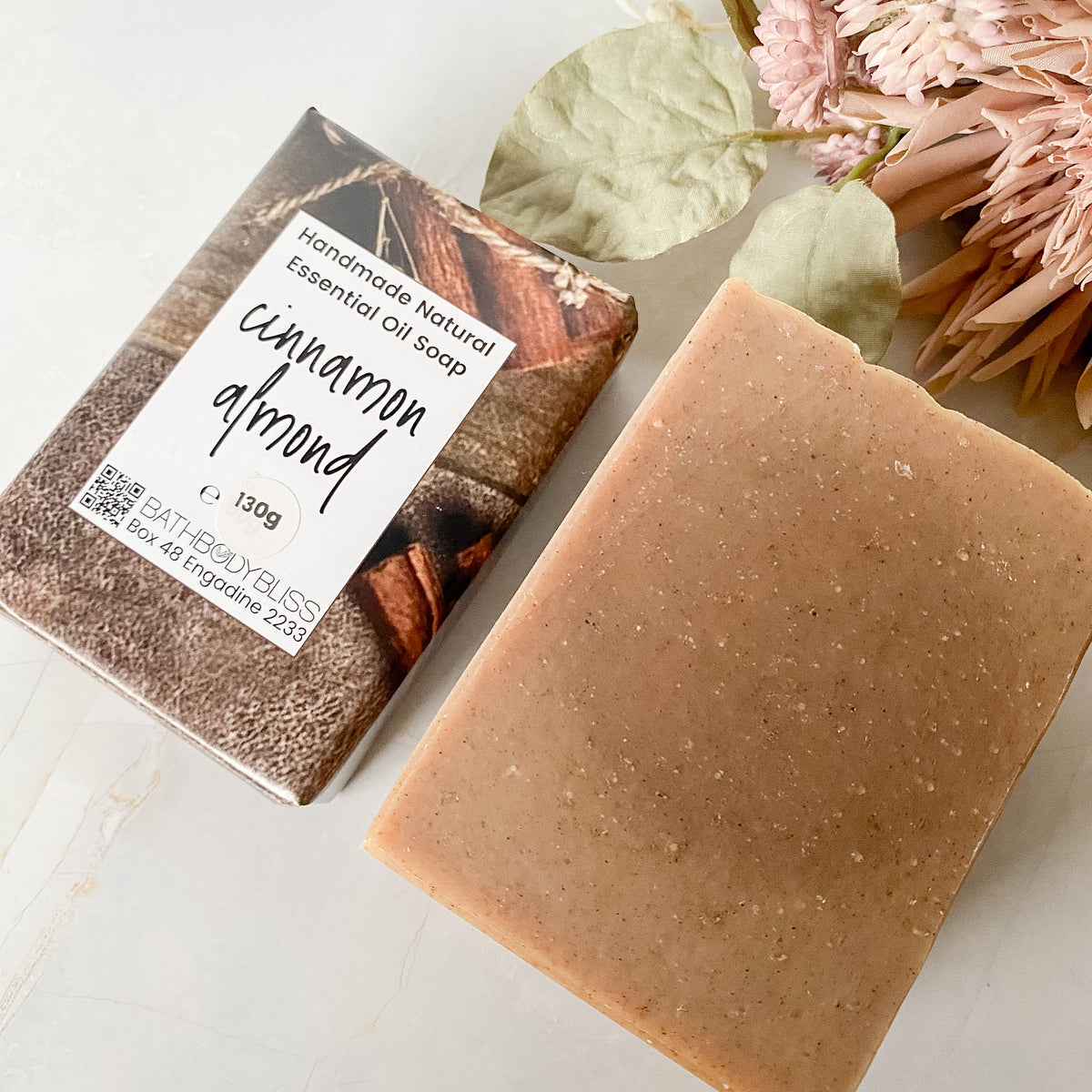 A bar of cinnamon almond essential oil soap shown next to its paper packaging, styled with dried leaves and pink flowers on a light marble surface.