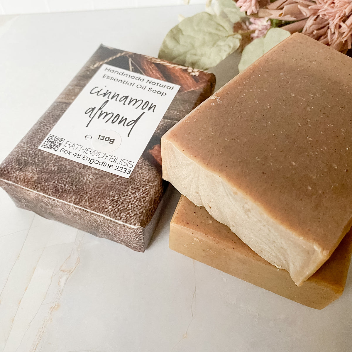 Three bars of handmade natural essential oil soap, one wrapped in brown paper packaging labeled 'cinnamon almond' and two unwrapped showing a smooth light brown surface, styled on a marble surface with dried flowers.