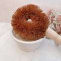 A brown natural coconut fibre dish and pot scrubbing brush with a wooden handle, resting in a white bowl.