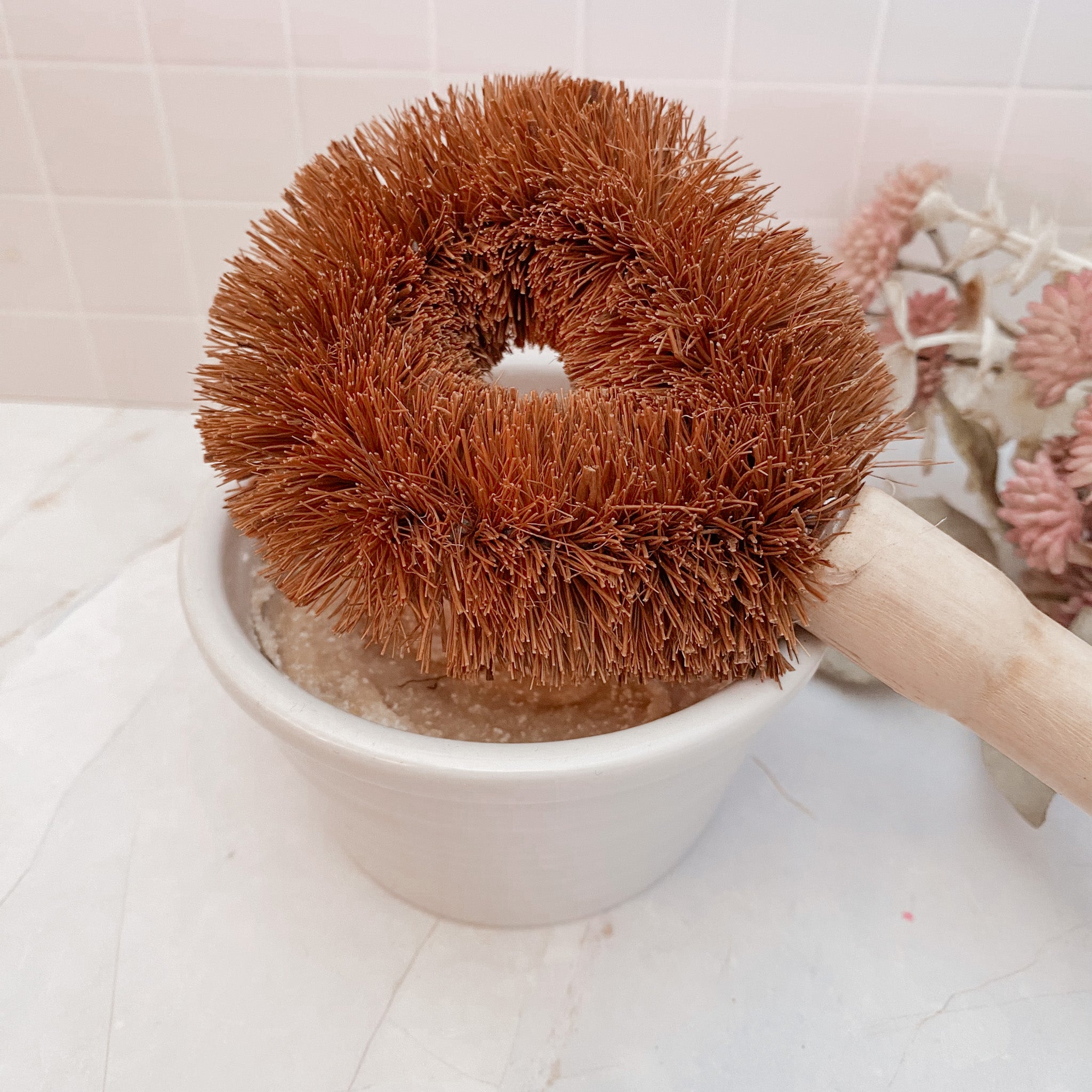 A brown natural coconut fibre dish and pot scrubbing brush with a wooden handle, resting in a white bowl.