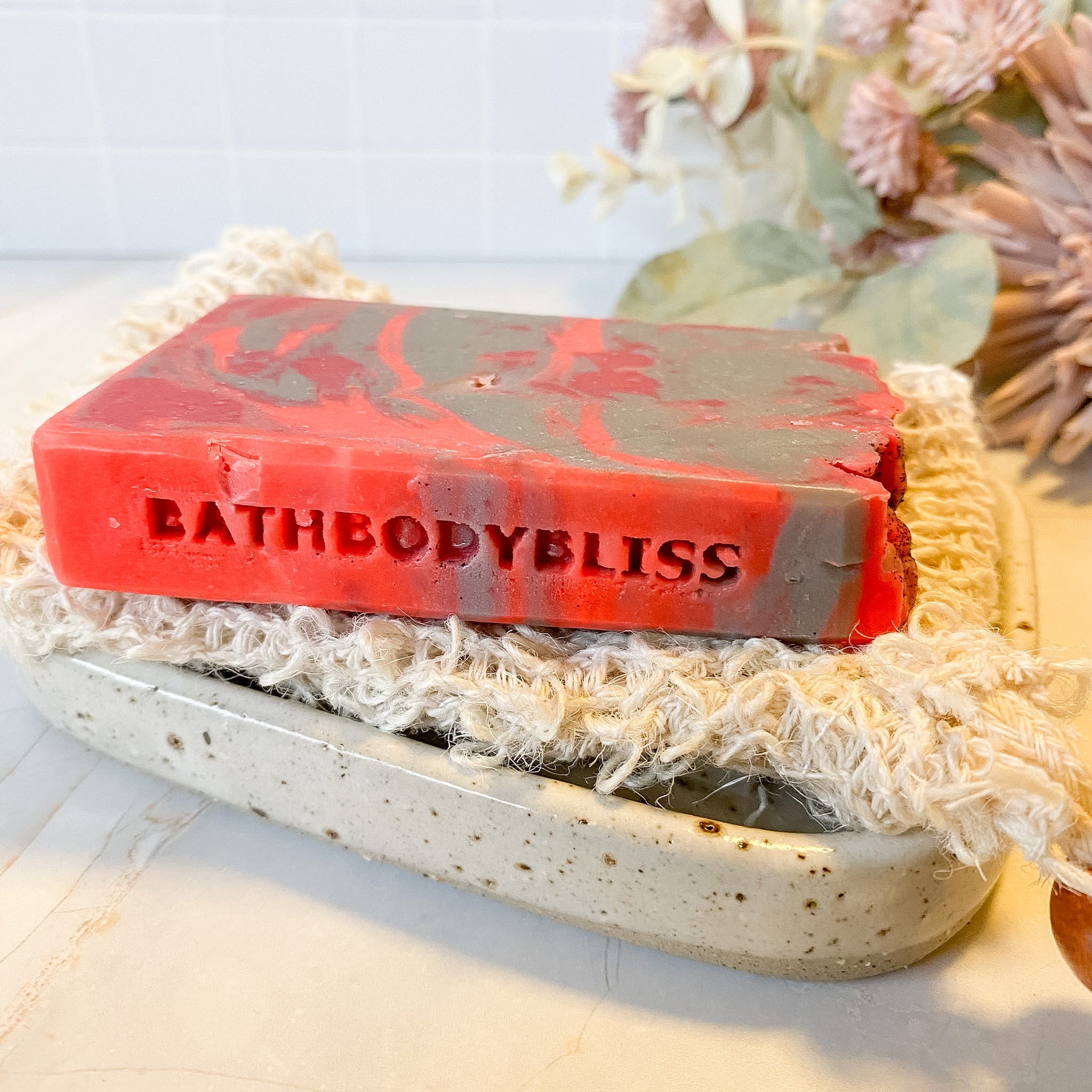 Red and gray soap bar with 'BathBodyBliss' branding on a textured soap dish.