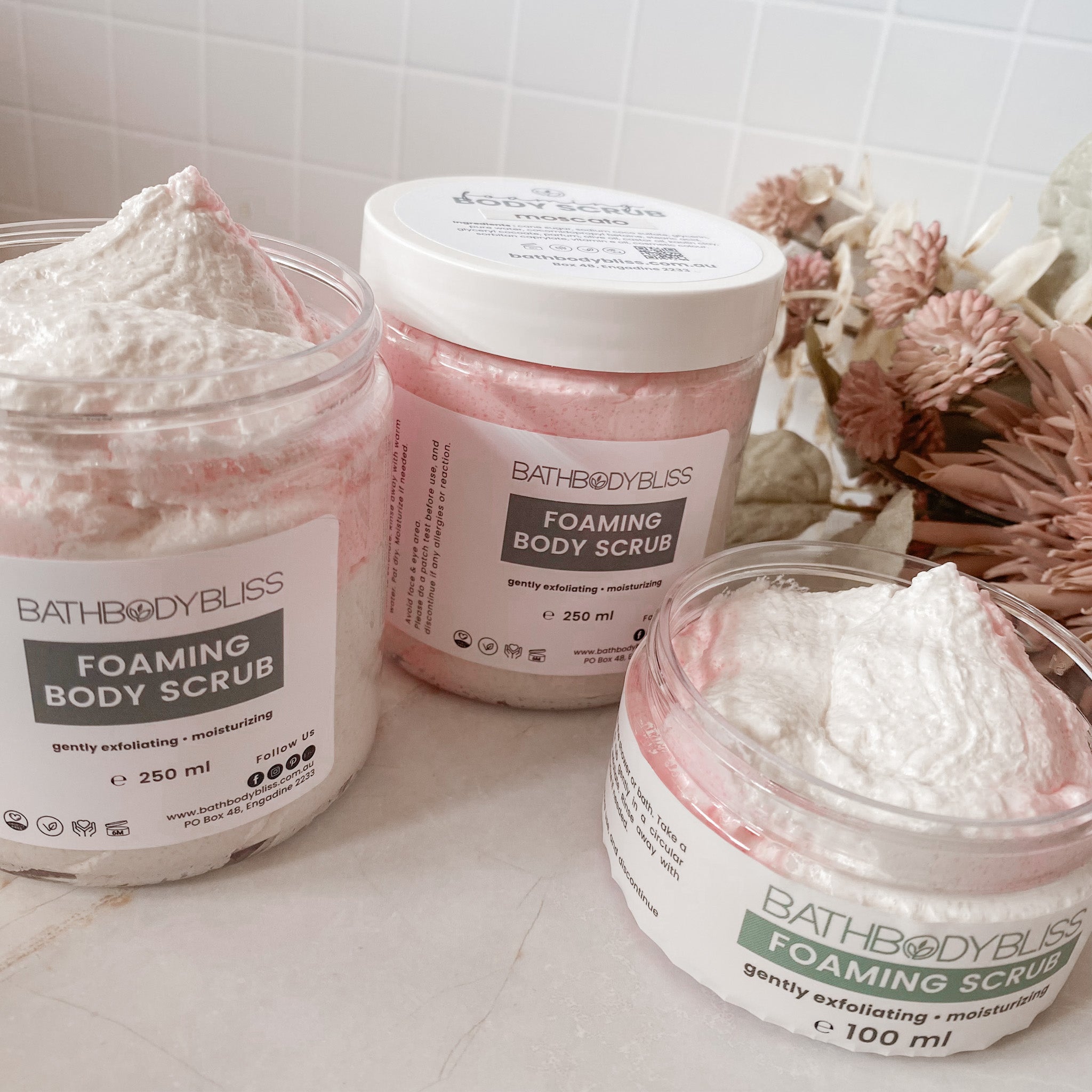 Three jars of Bath Body Bliss foaming body scrub on a light surface with a floral arrangement in the background.