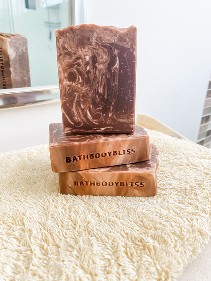 Three bars of BathBodyBliss soap stacked on a fluffy white surface with a blurred background.