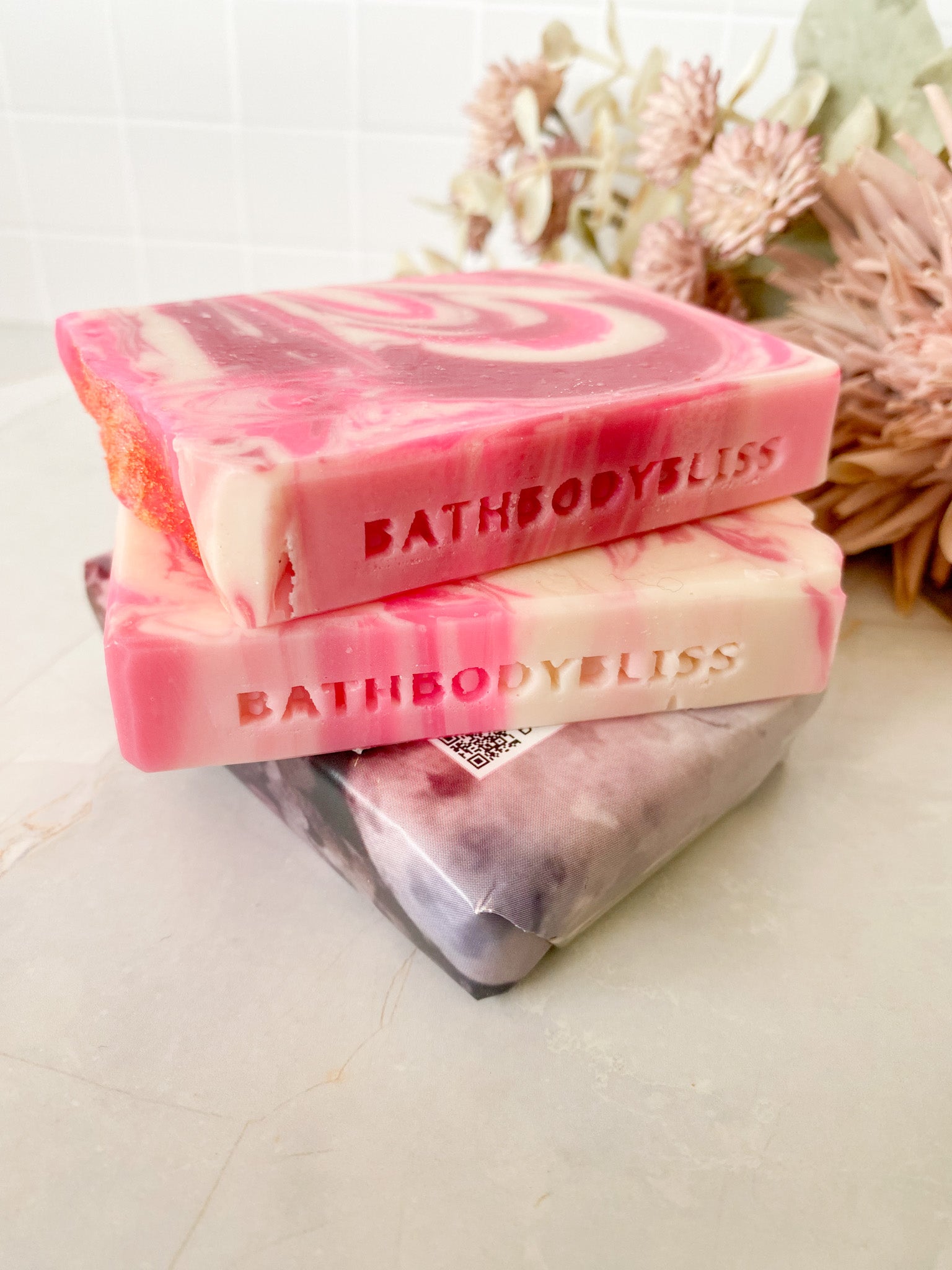 Handmade soap bars with a swirl pattern in shades of pink and white, branded 'BATHBODYBLISS', displayed alongside decorative dried flowers.