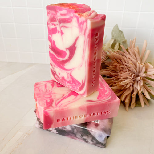 A stack of pink and white marbled soap bars with the text 'BATHBODYBLISS' visible on one of the bars, placed on a white surface with a flower in the background.