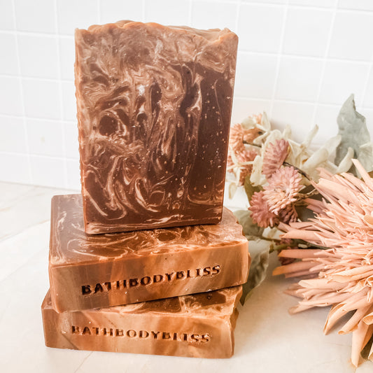 Three bars of BathBodyBliss soap stacked on a white surface with flowers in the background.