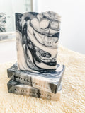 A bar of handmade vegan soap with a black and gray marbled pattern, placed on a textured surface.