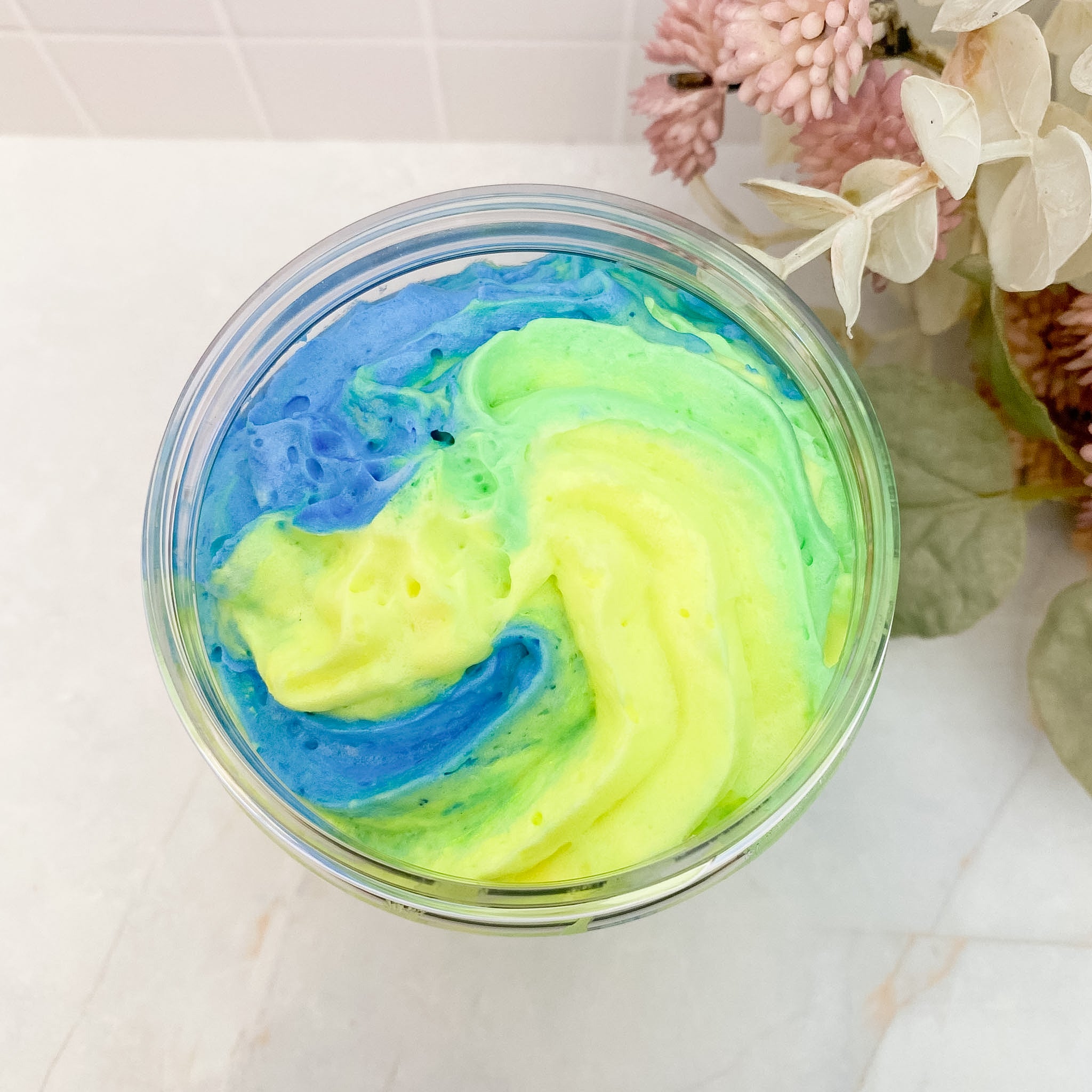 Swirl of blue and green whipped soap in a clear container on a white surface with flowers in the background.