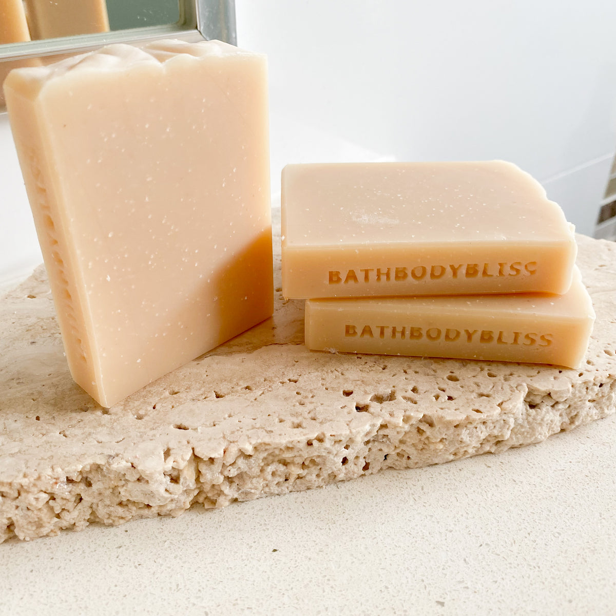 Three bars of handmade goatsmilk soap with 'BATHBODYBLISS' stamped on the sides, placed on a natural-colored stone surface.