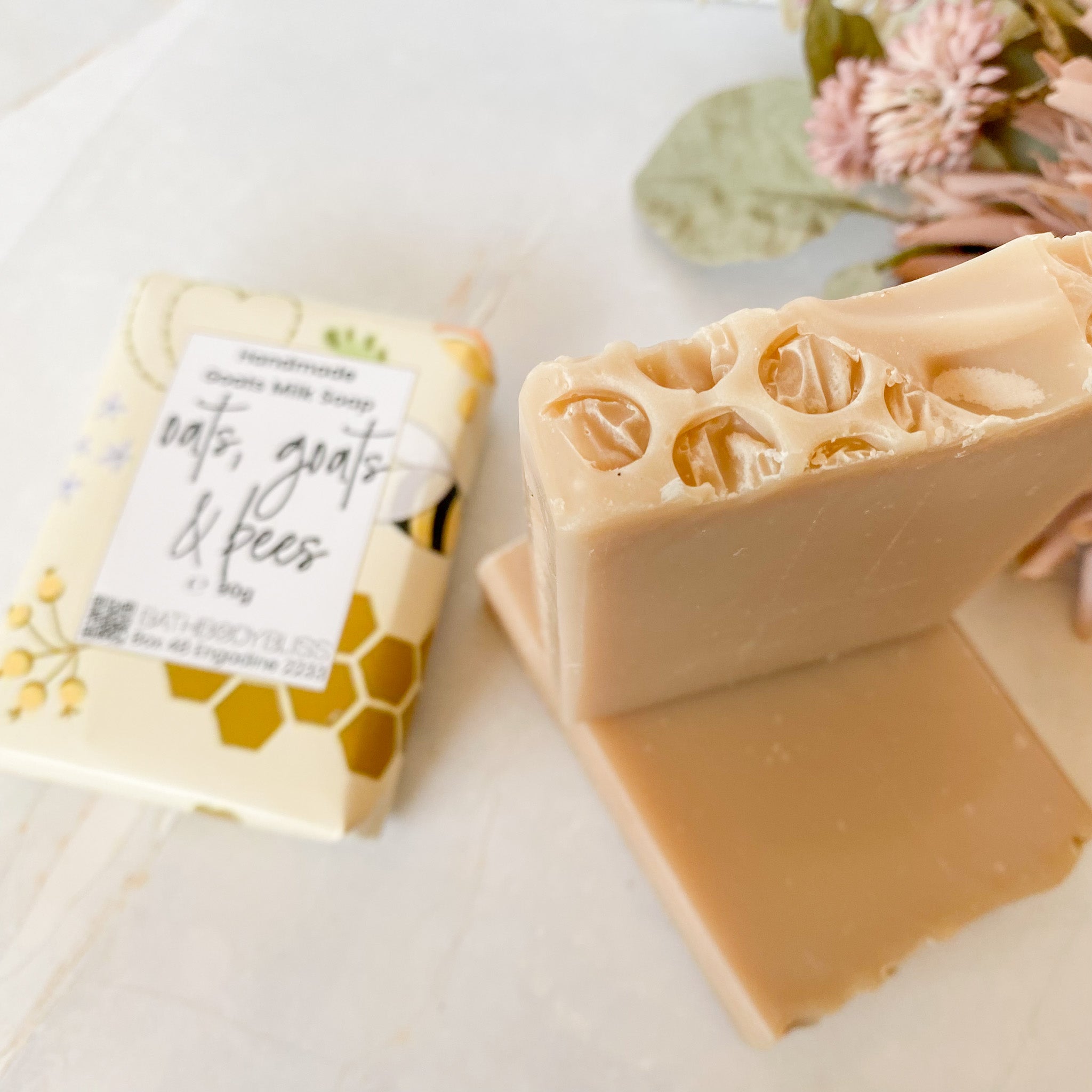 A handmade unscented goat milk soap bar with a honeycomb texture, shown next to its cream-colored paper packaging labeled 'oats, goats & bees' on a light marble surface.