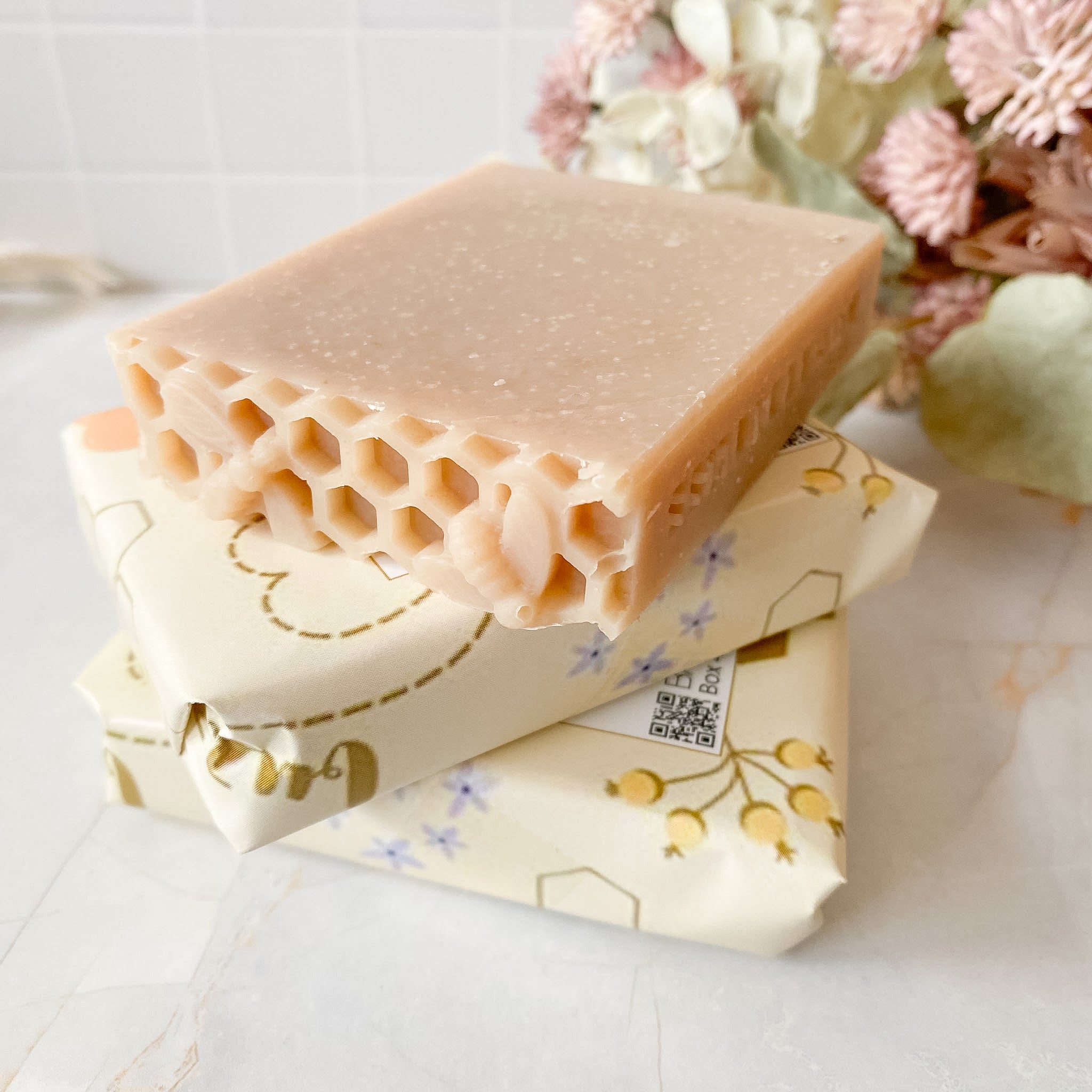 A rectangular bar of honeycomb-textured soap in a warm peach-beige color, resting on top of two cream-colored wrapped bars with gold and blue floral designs. The background is a soft-focus white tiled wall and marble surface with dried flowers.