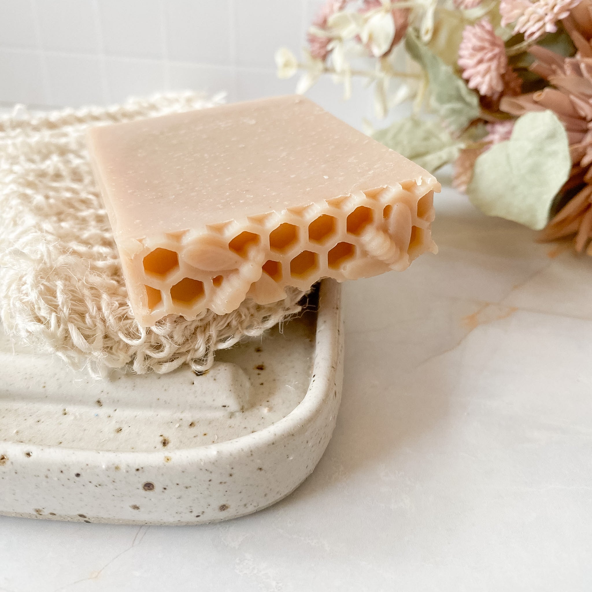 A rectangular bar of pale peach-beige honeycomb soap resting on a white textured washcloth in a speckled ceramic soap dish, with dried flowers in the background.