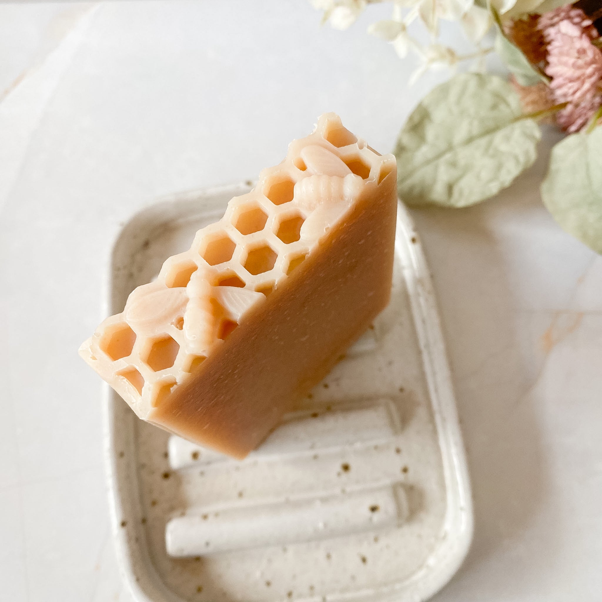 A rectangular bar of honey-colored soap with a raised honeycomb pattern on one end, resting on a speckled white soap dish with dried flowers in the background.