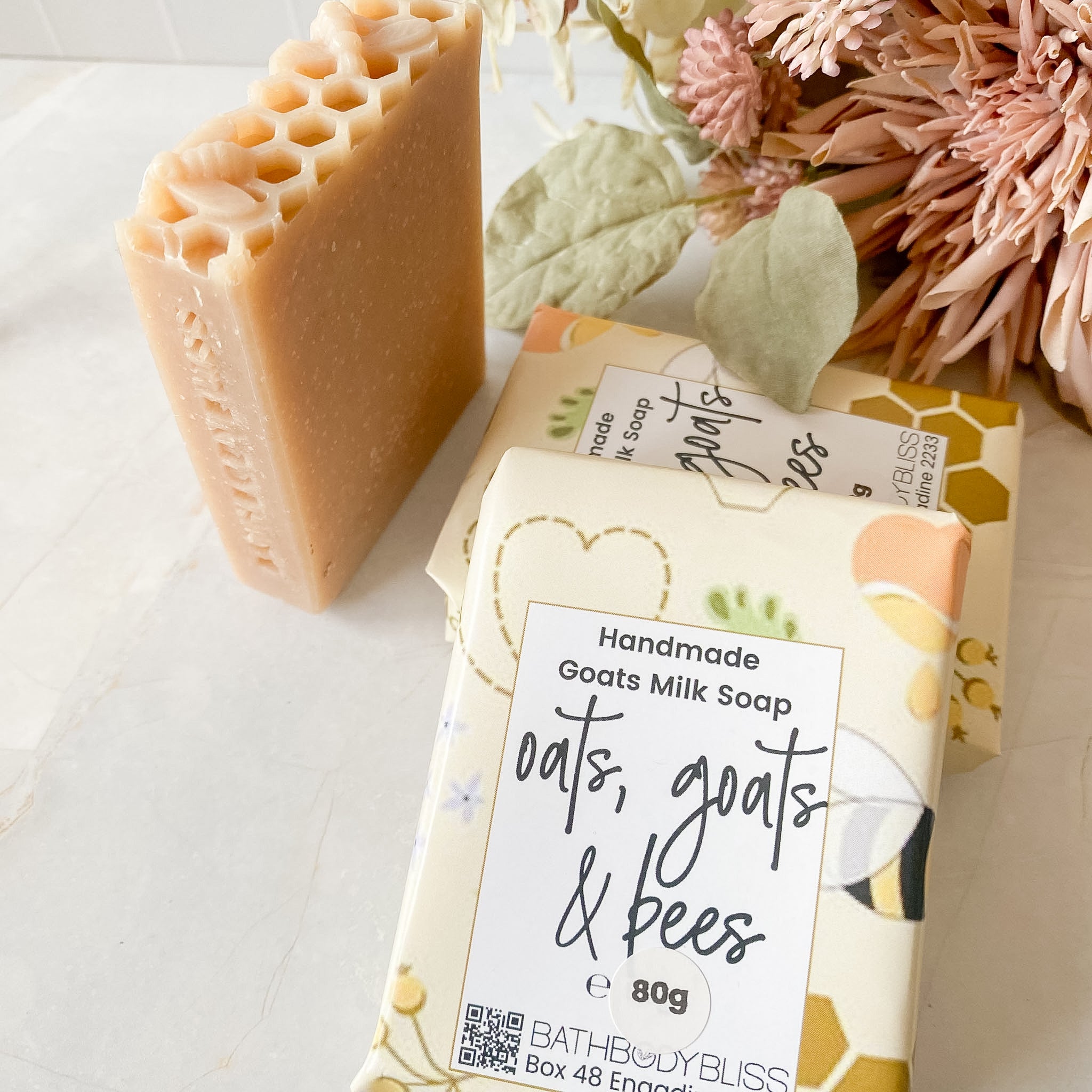 Handmade goat's milk soap bars from Bath Body Bliss Oats, Goats & Bees, shown with their illustrated packaging on a light marble surface next to dried flowers.