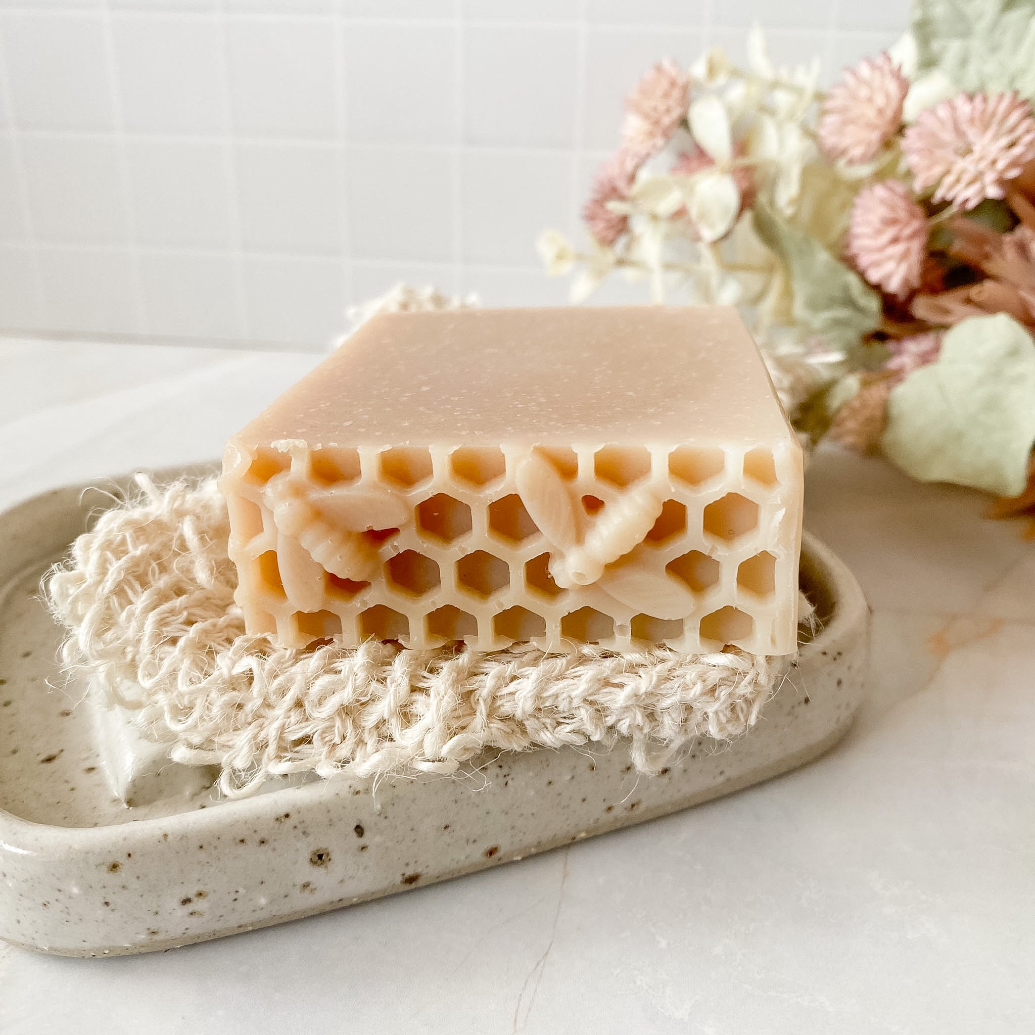 soap bar with honeycomb and bee impression sitting on a soap dish and washcloth with floral decor in background