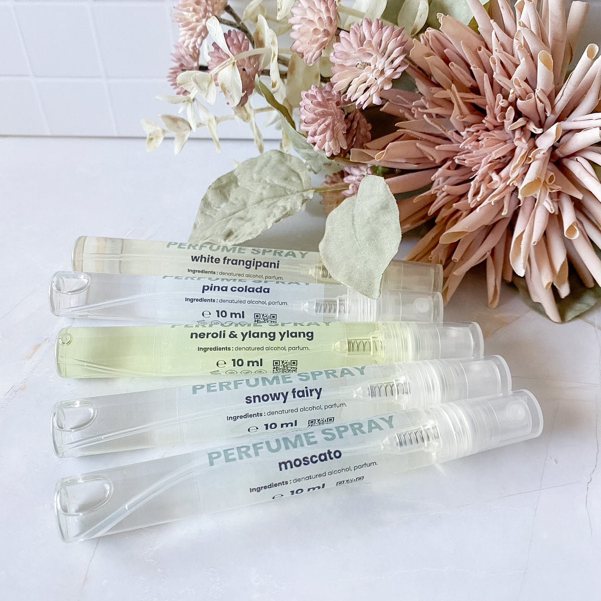 Five clear perfume spray vials with printed labels are arranged diagonally on a white marble surface next to dried pink flowers. The visible labels include white frangipani, pina colada, neroli & ylang ylang, snowy fairy, and moscato.