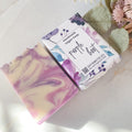 Handmade vegan soap bar with packaging labeled 'Purple Hoot' on a light surface with flowers.
