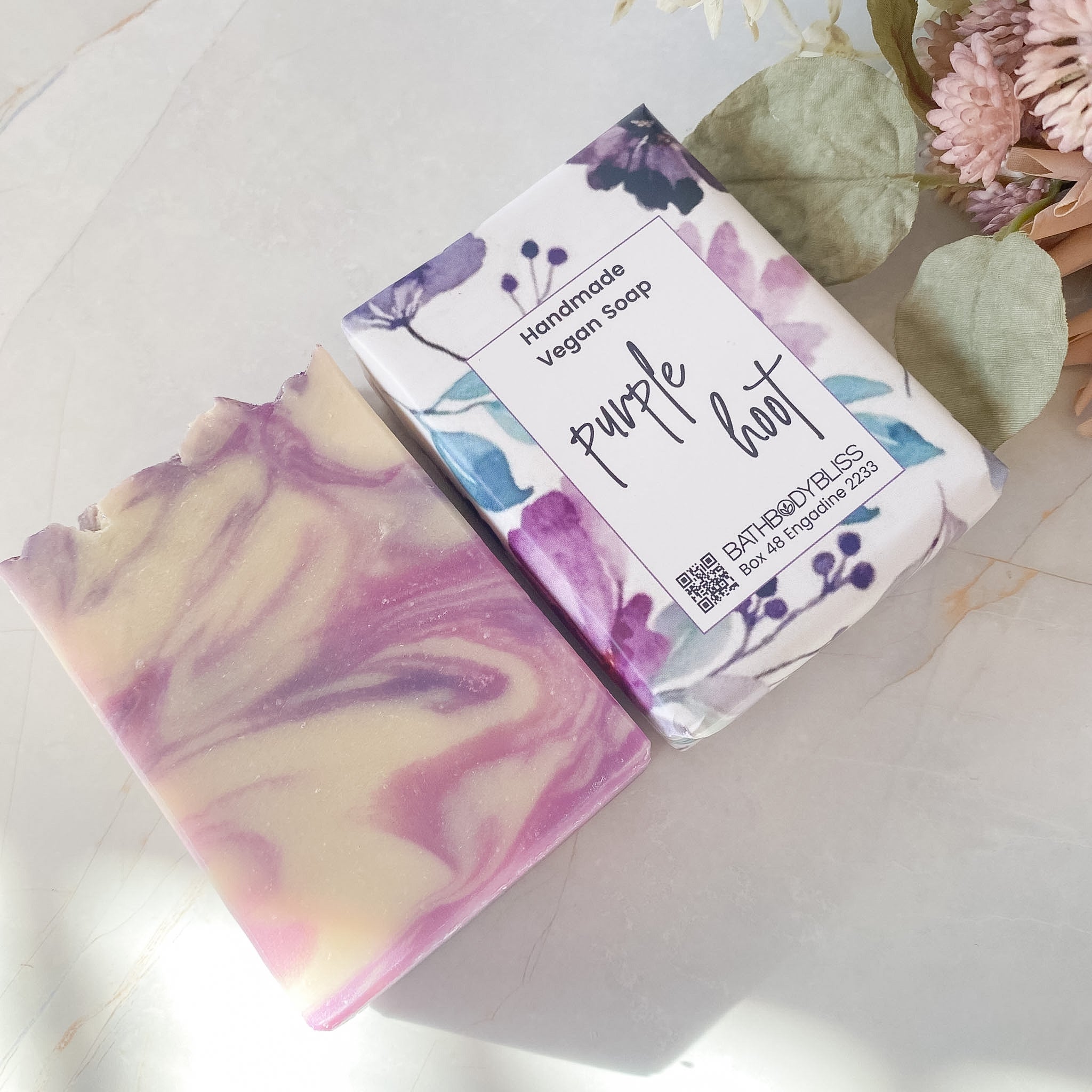 Handmade vegan soap bar with packaging labeled 'Purple Hoot' on a light surface with flowers.