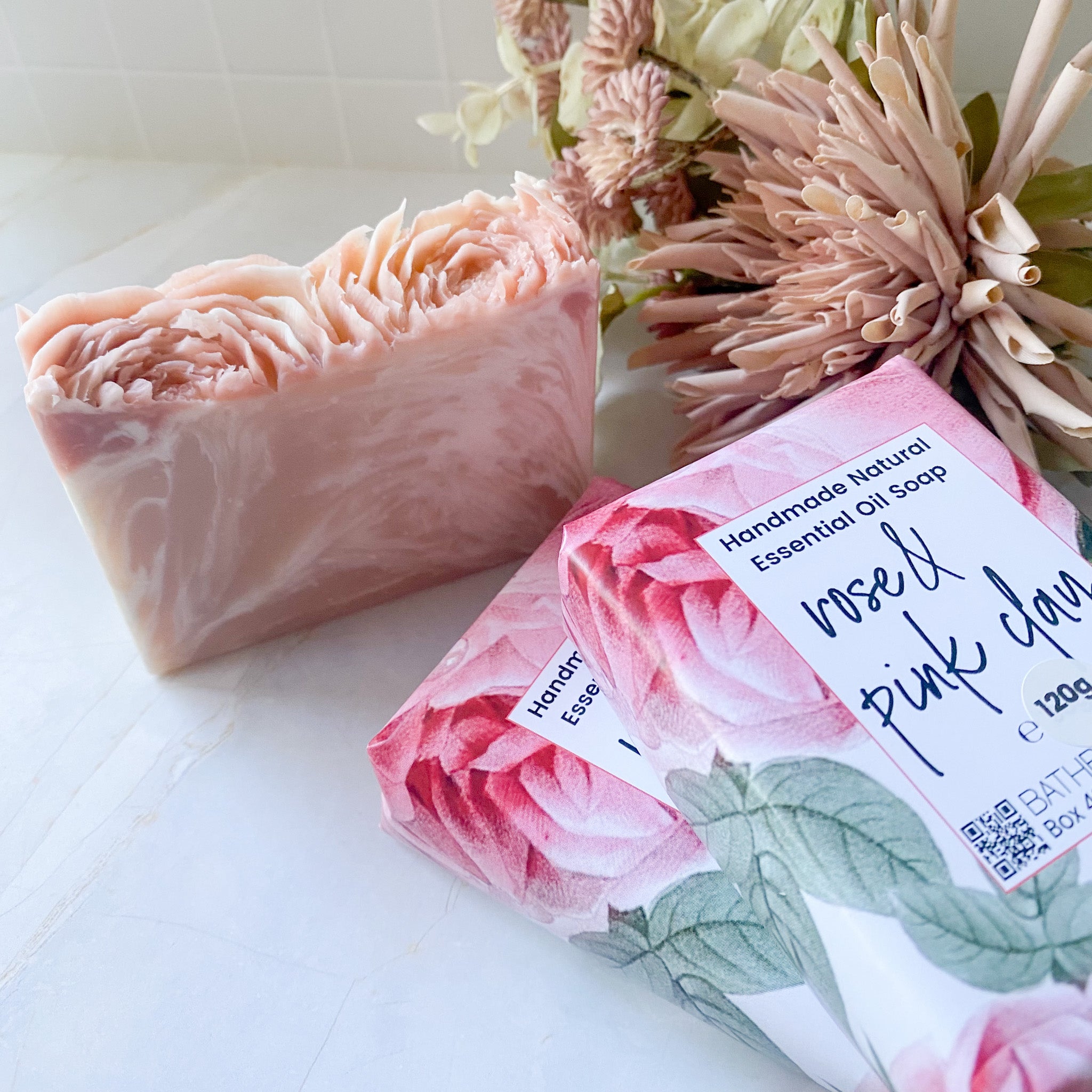 Handmade natural essential oil soap with a pink and green label, placed on a white surface with flowers in the background.