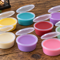 Open round clearplastic containers with lids containing coloured wax melts, arranged on a wooden tabletop.