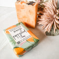 orange poppyseed soap bar in bath body bliss packaging and a standing unwrapped soap bar with orange swirls and poppyseeds on top next to it