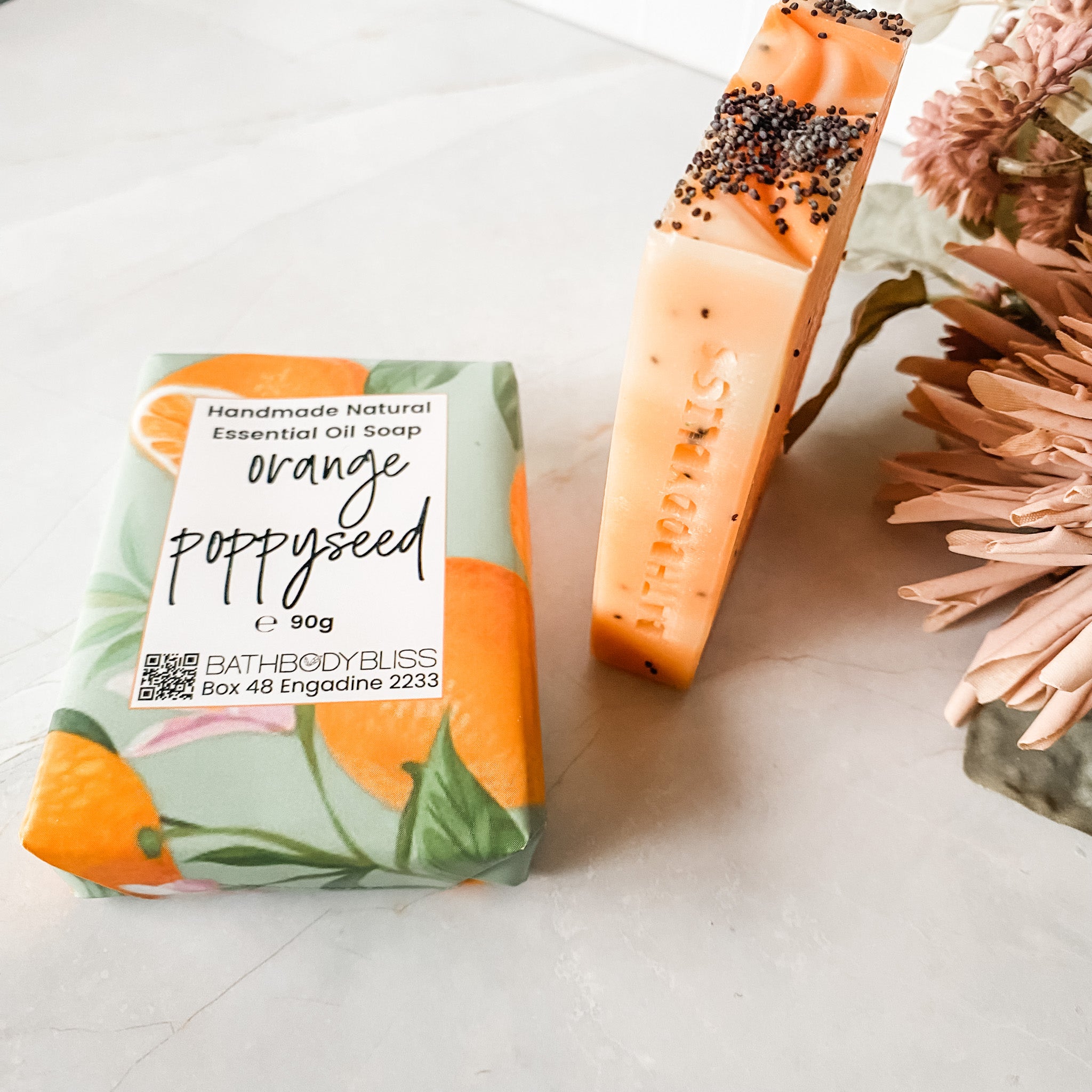 orange and poppyseed soap bar in bath body bliss packaging and unwrapped soap bar with orange swirls and poppyseeds standing beside it
