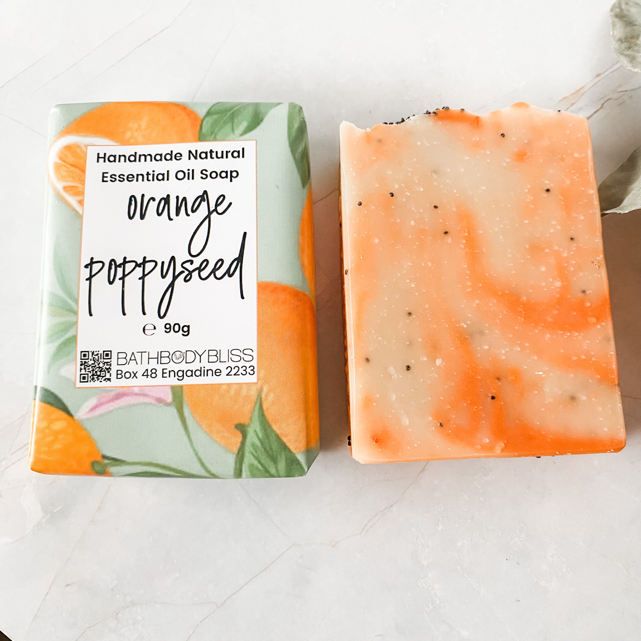 orange and cream swirled soap bar with poppyseeds in bathbodybliss branded paper wrap and unwrapped soap bar beside it on a white background