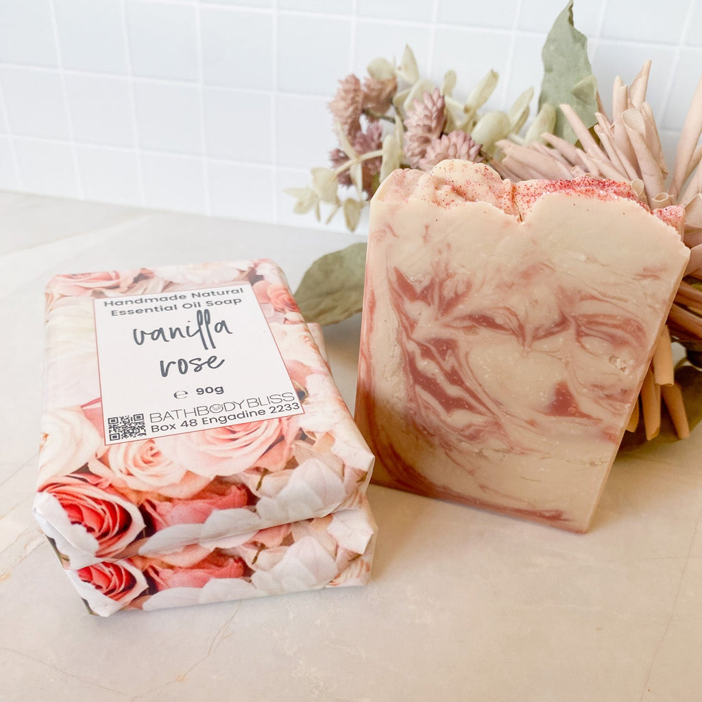 Three bars of soap, two wrapped in floral paper and labeled 'Vanilla Rose', on a light surface with a decorative plant in the background.