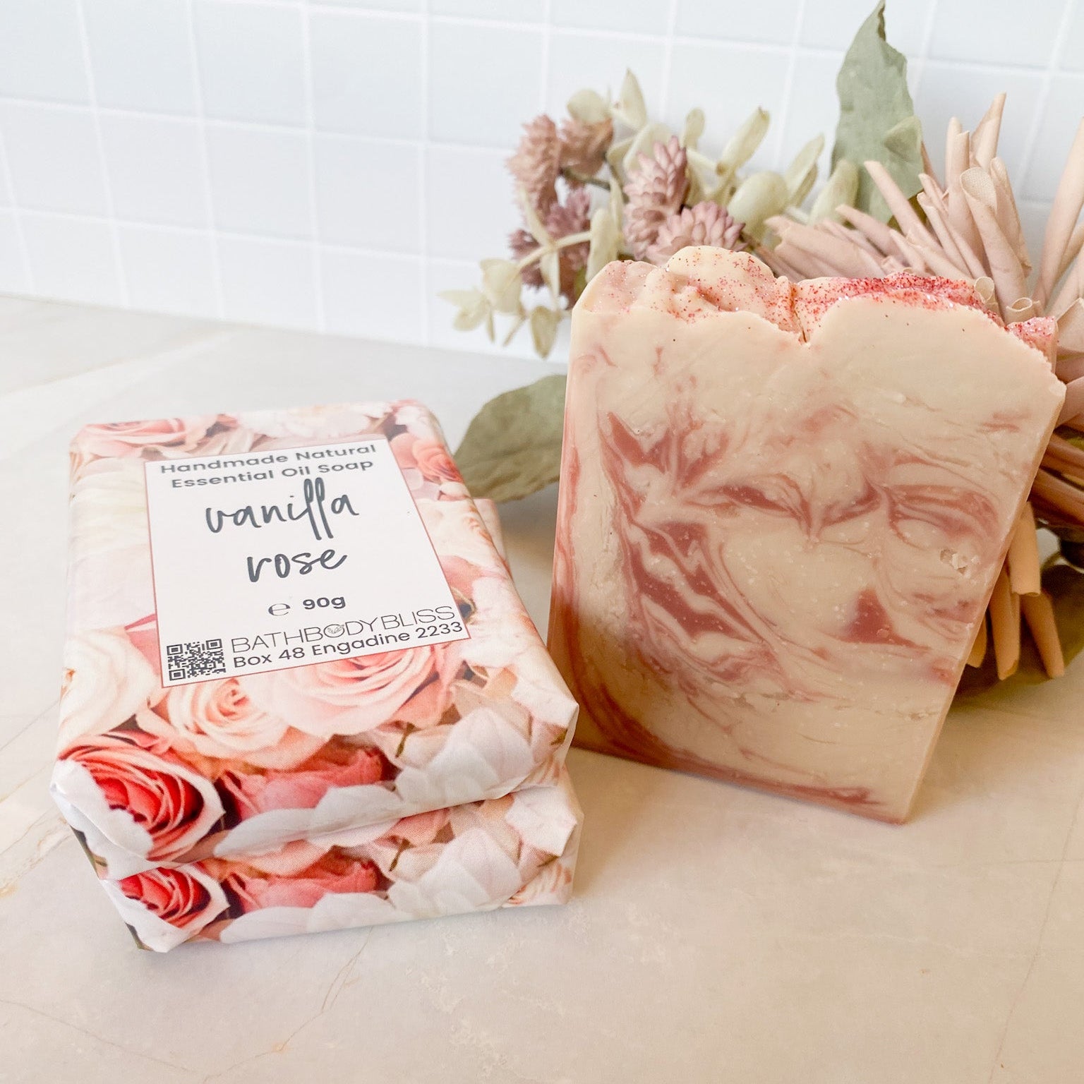Three bars of soap, two wrapped in floral paper and labeled 'Vanilla Rose', on a light surface with a decorative plant in the background.