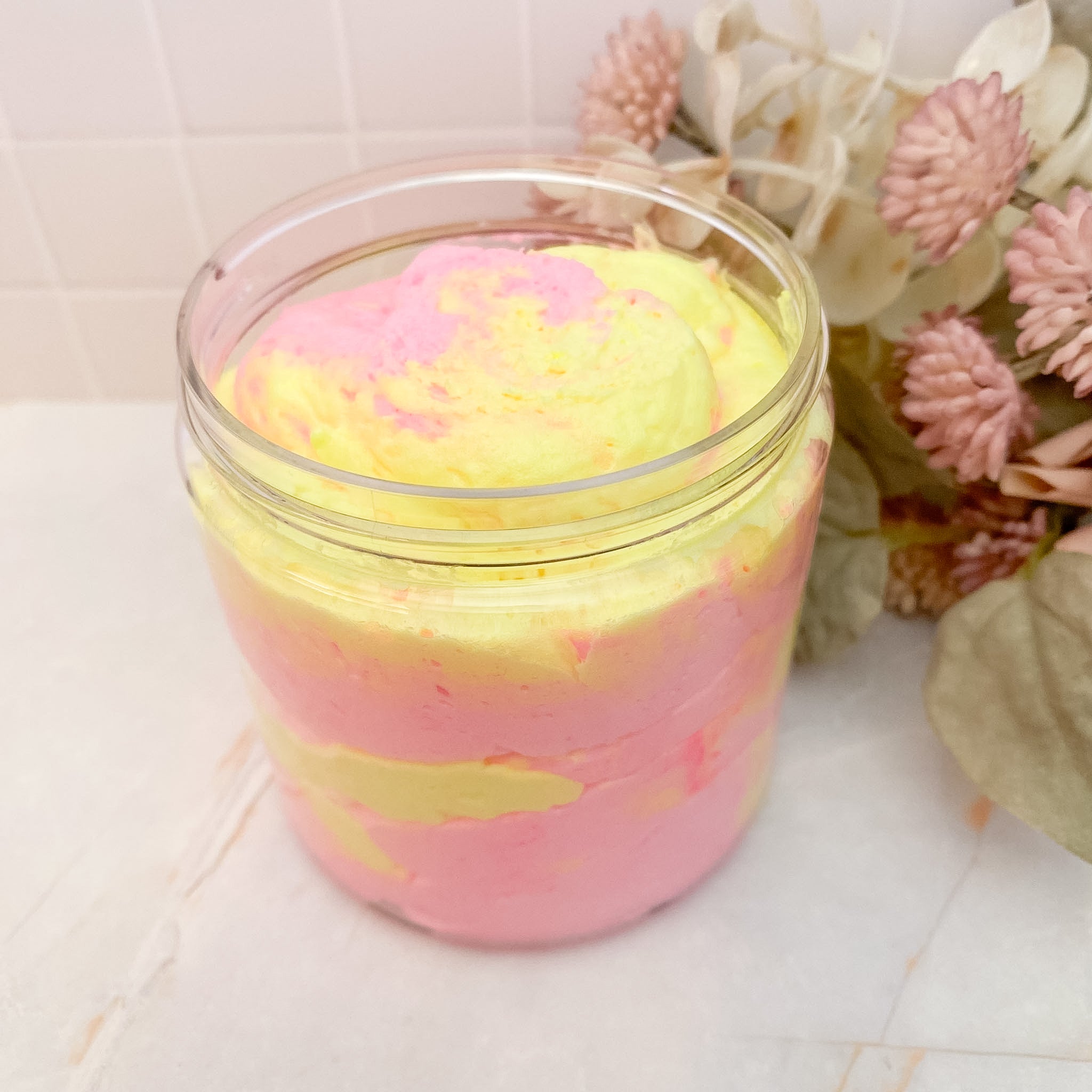 Jar of pink and yellow whipped soapwith decorative flowers in the background