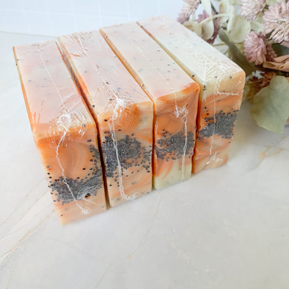 four shrink wrapped handmade soap bars with orange swirls and poppyseed decoration, on a light marble background with floral decor