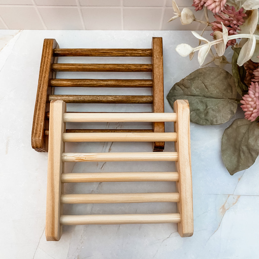 Two wooden soap dishes on a light marble background with decorative plants in the background.