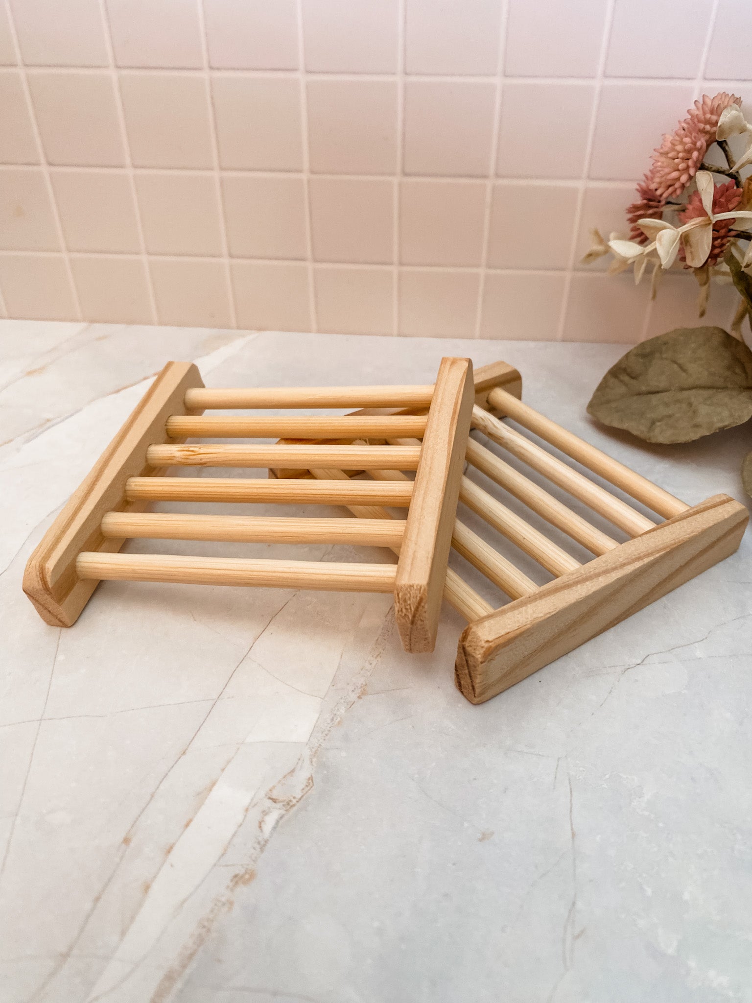 A square bamboo soap dish placed on a marble surface, with a few leaves in the background.