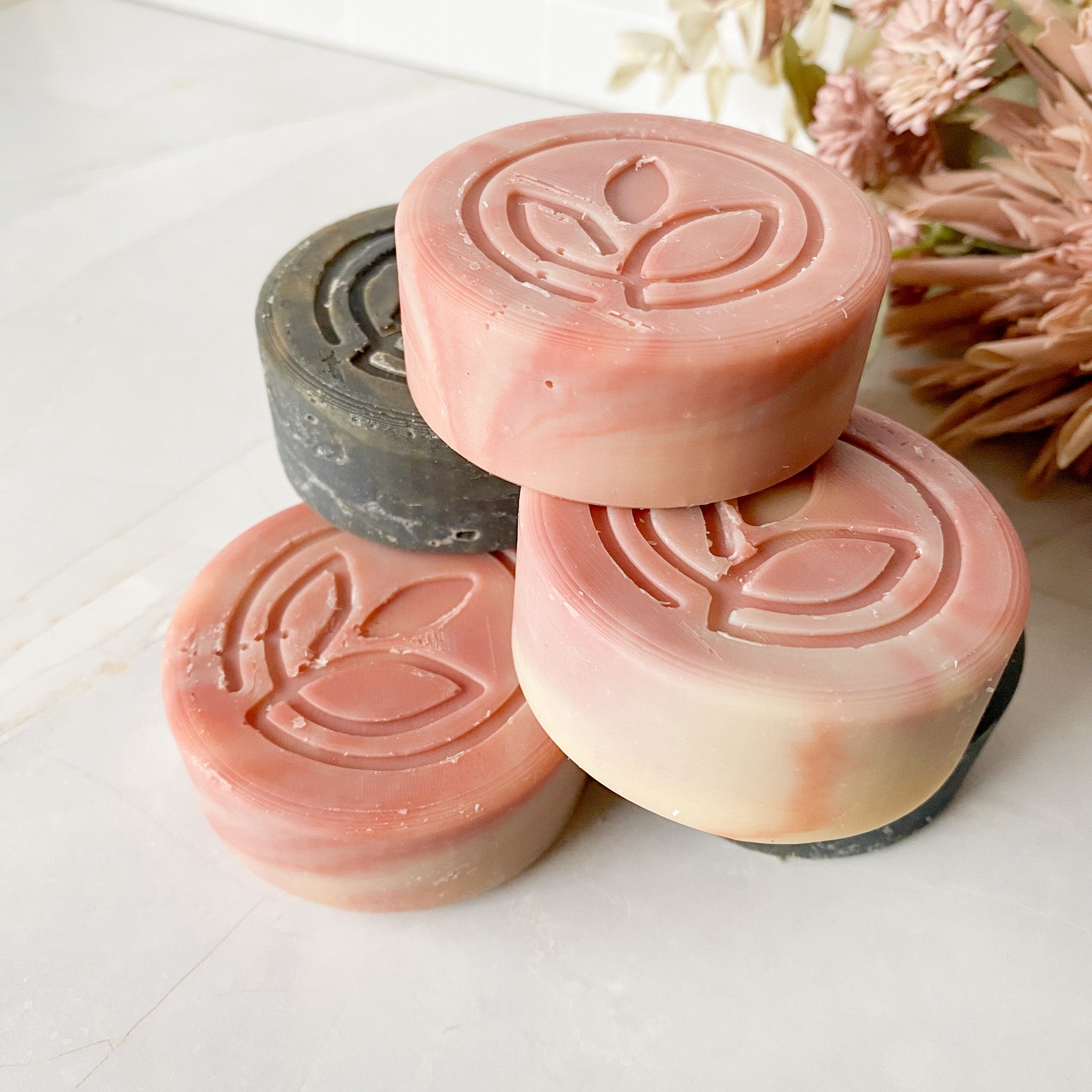 Five round soap bars with a leaf design on a light surface, with dried flowers in the background.