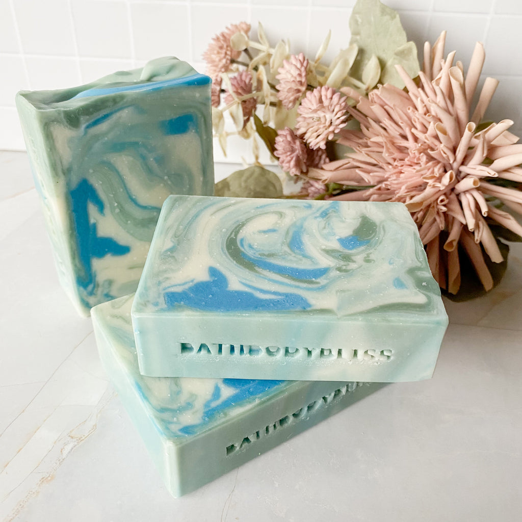 Three swirled soap bars with 'Bath Body Bliss Bliss' branding on a white surface with flowers in the background.