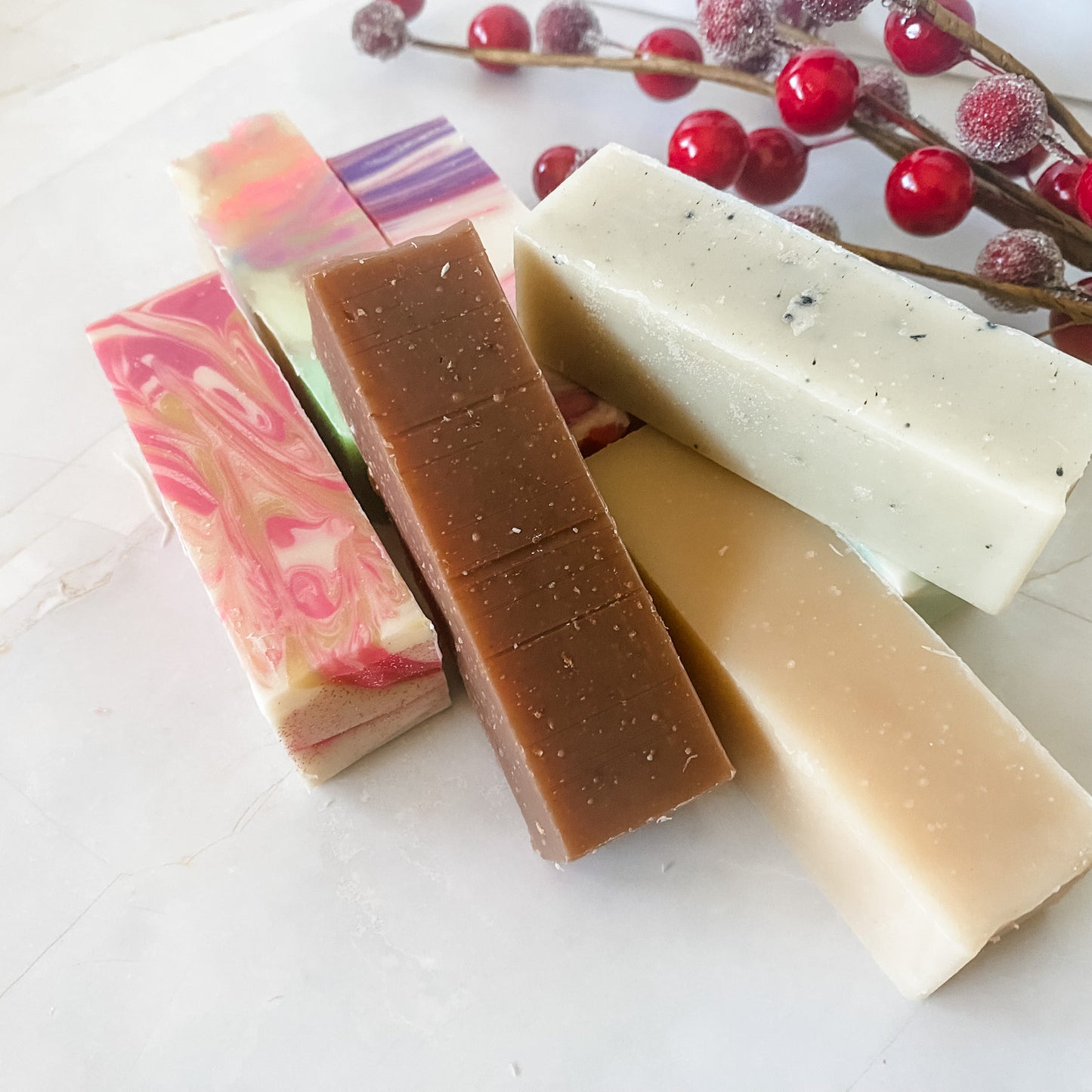 Six bars of soap in different colors on a white surface with red berries in the background